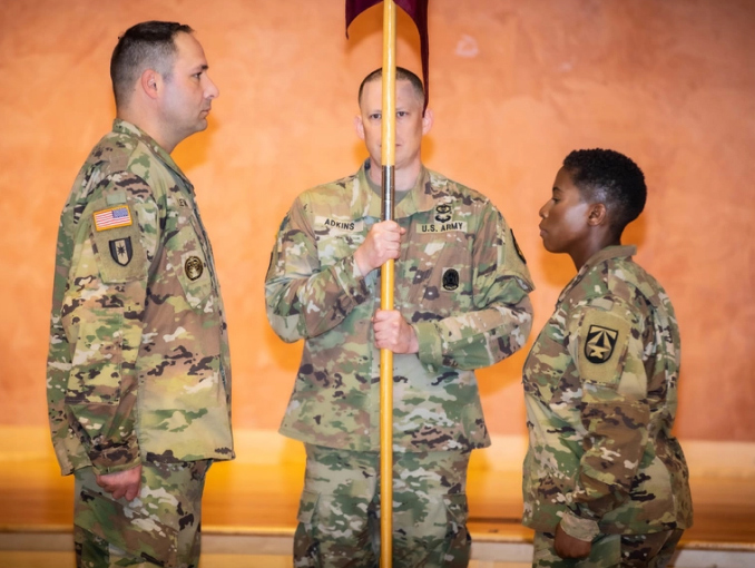 A female and two male Soldiers in combat uniform stand in position to pass a flag during a ceremony