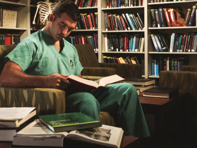 Medical student in scrubs reading a textbook in a library