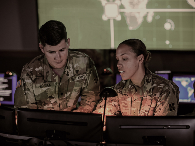 A female Soldier training a male Soldier in front of two computer monitors