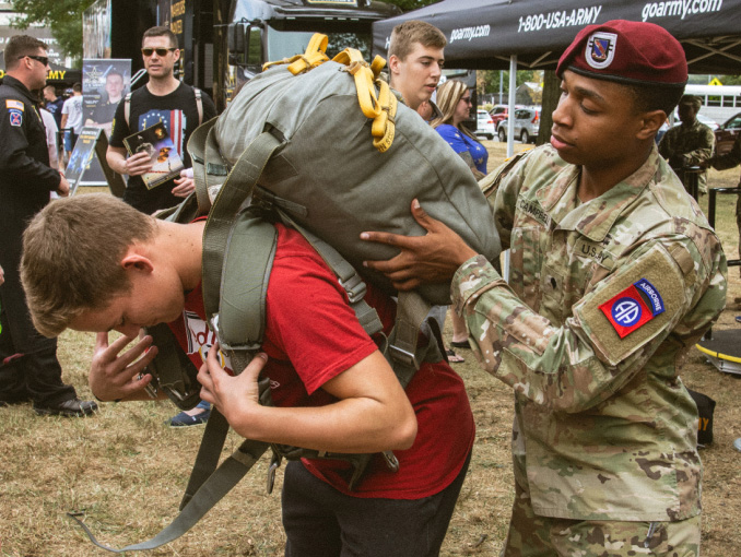 A Soldier assisting a civilian teenage boy put on an Army rucksack at a recruiting event