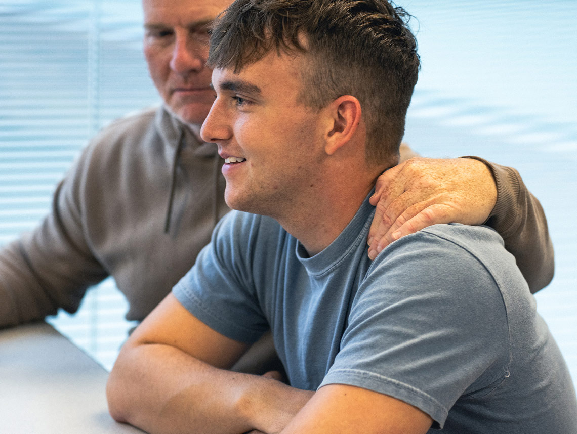 A male enlisted recruit wearing a light blue t-shirt, sitting down and staring into the distance with another person’s arm wrapped around his shoulder