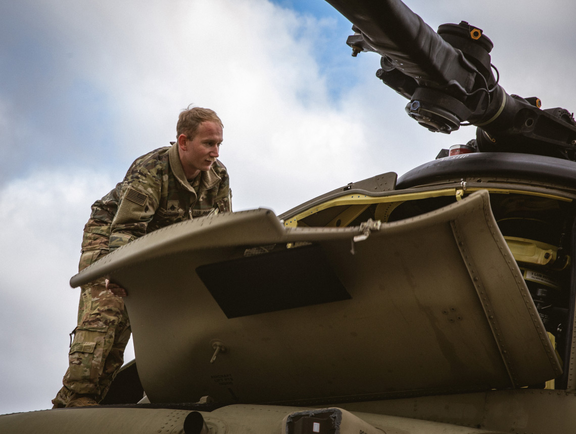 Army Sergeant standing on top of a helicopter hunched over with blue skies and scattered clouds in the background.