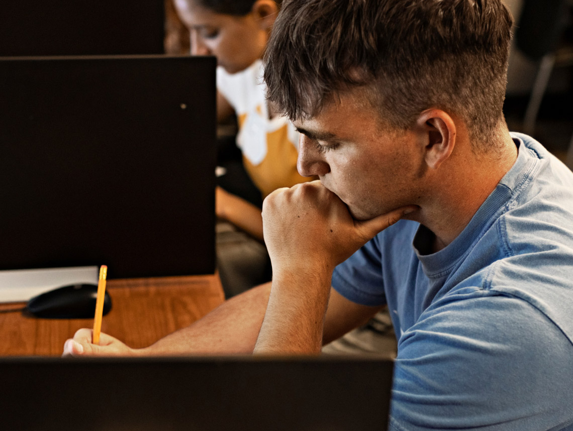 Un recluta vestido con una camiseta azul claro, sentado en un escritorio café y escribiendo con un lápiz