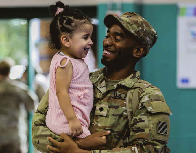 Un Soldado en uniforme de combate sonríe sosteniendo a su hija pequeña