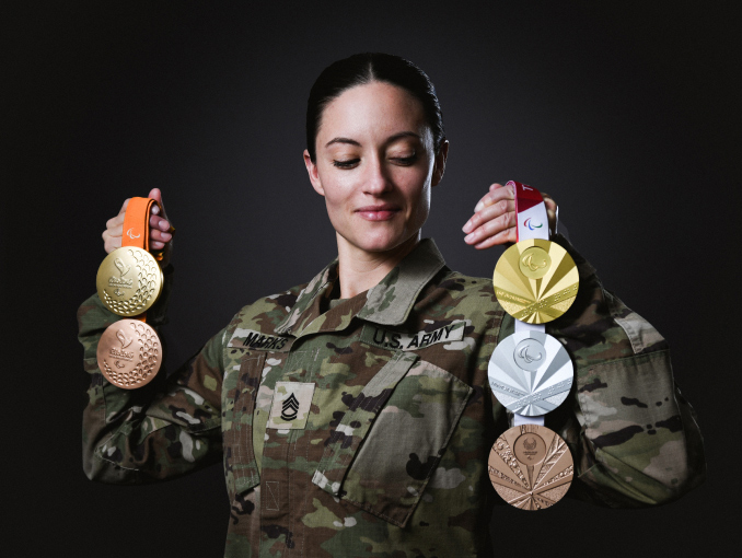A female Soldier in combat uniform holding medals from various Olympics