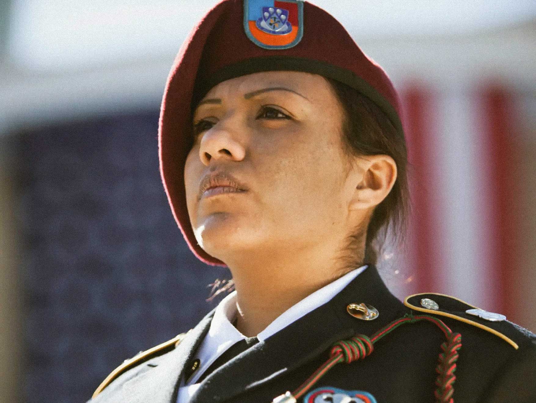 A female Specialist wearing Class A uniform standing outside during the day in front of the US flag