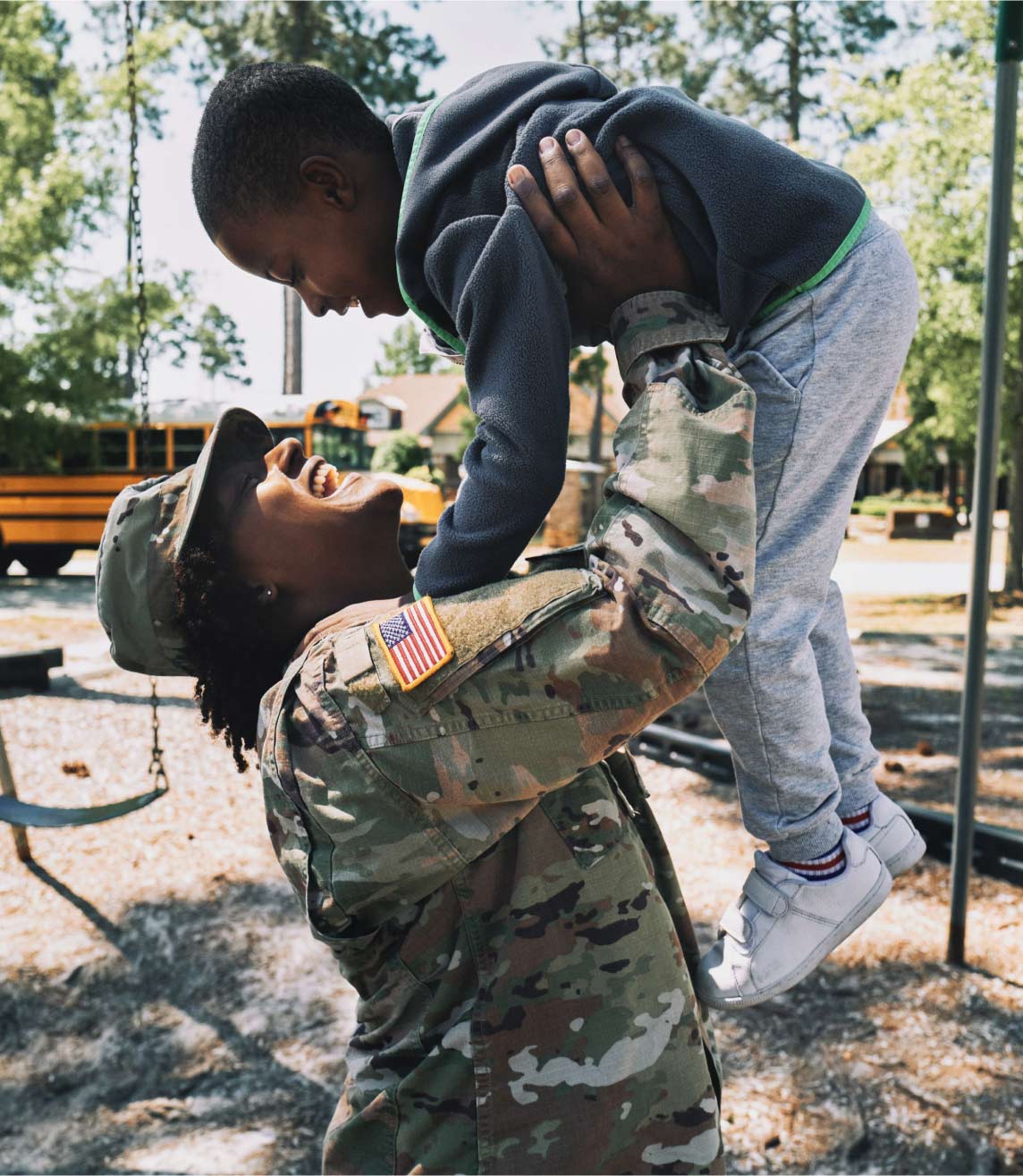 Una Soldado en uniforme de combate sostiene a su hijo en el aire en un patio de juegos