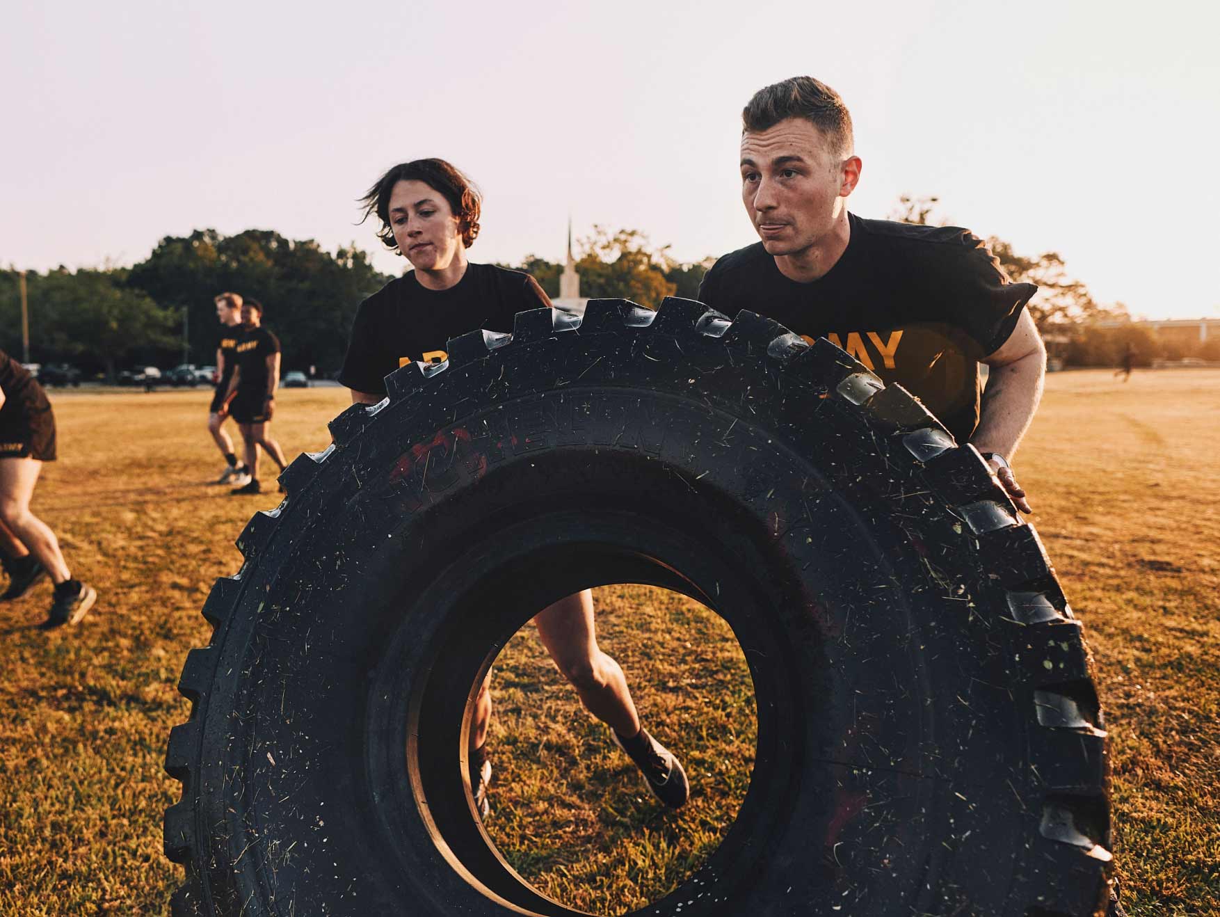 A male Soldier in an Army tee shirt pushing a tractor tire in a field as part of an exercise