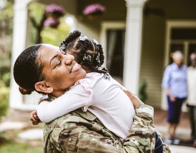 Una Soldado sonriendo y abrazando a su hija pequeña frente a su casa