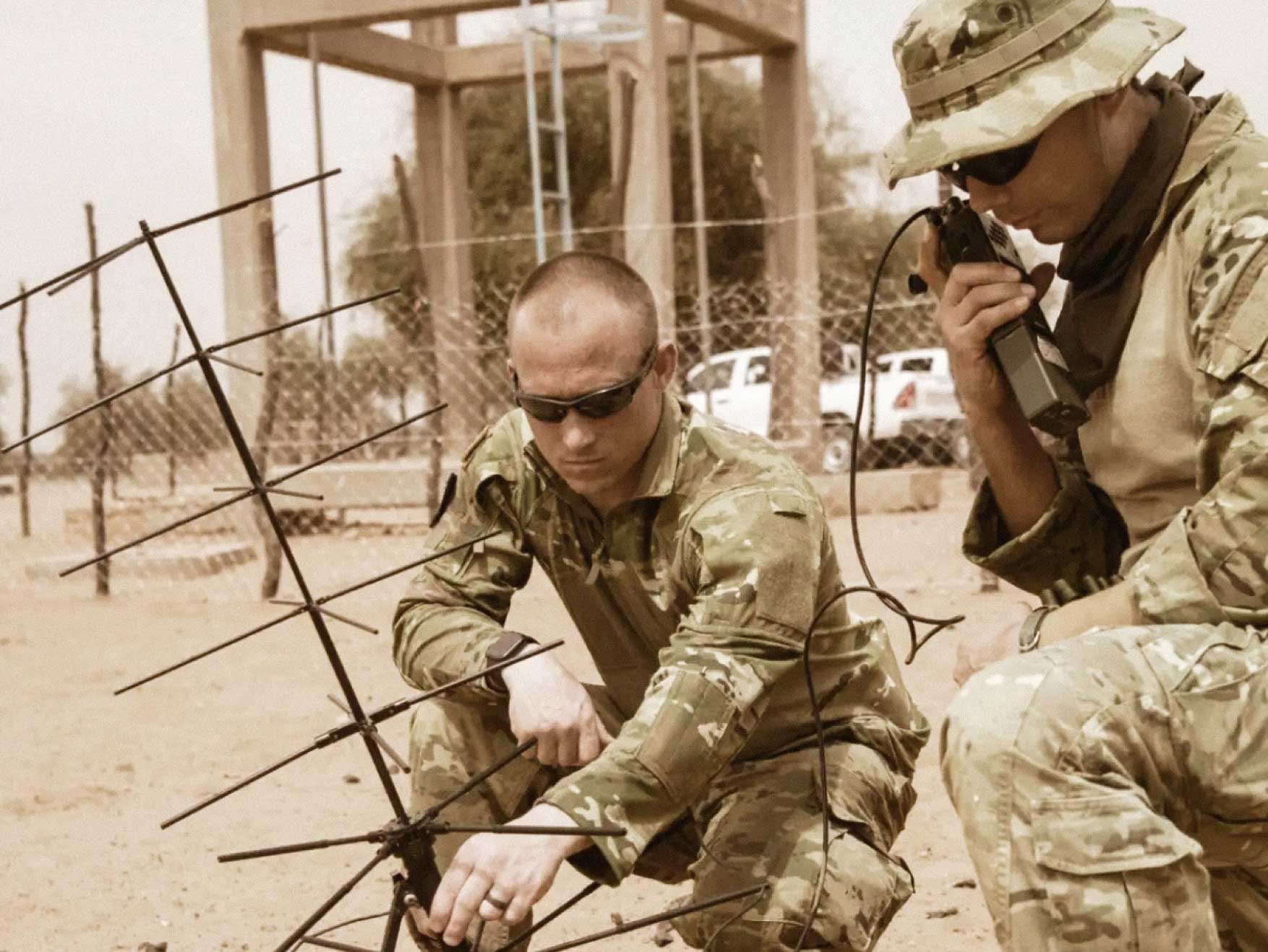 Dos Soldados de Psychological Operations arrodillados en la arena instalando tecnología de comunicaciones