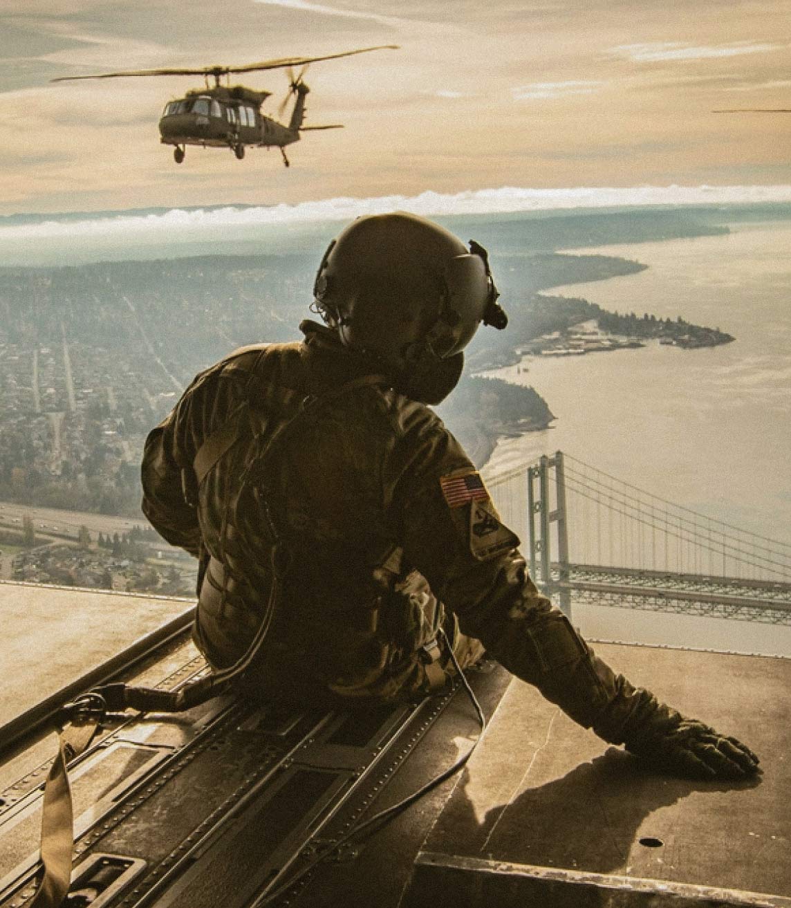 Un paracaidista sentado a bordo de un helicóptero en vuelo y a lo lejos dos helicópteros volando