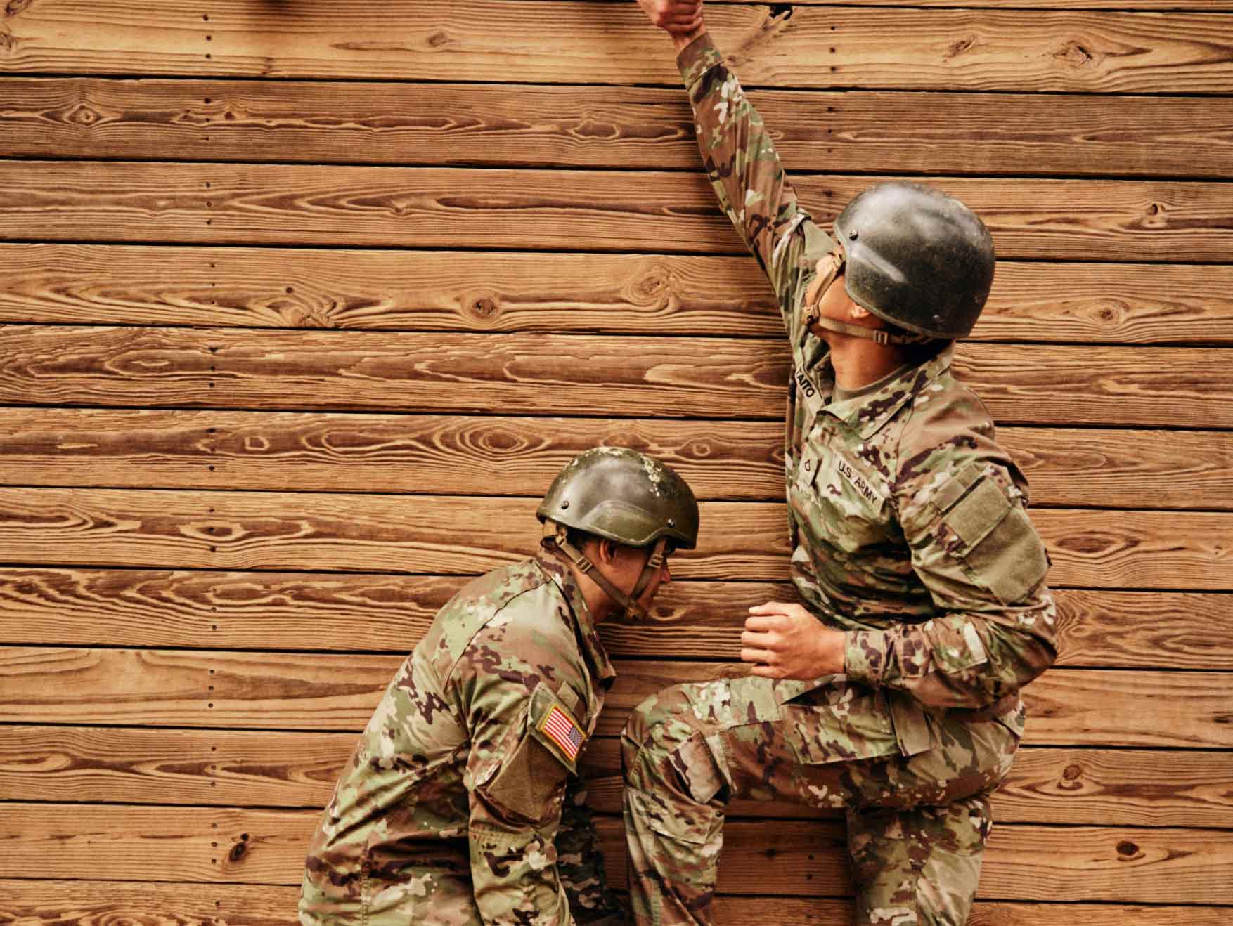 A Soldier bracing the foot of another Soldier to help him over a wooden obstacle