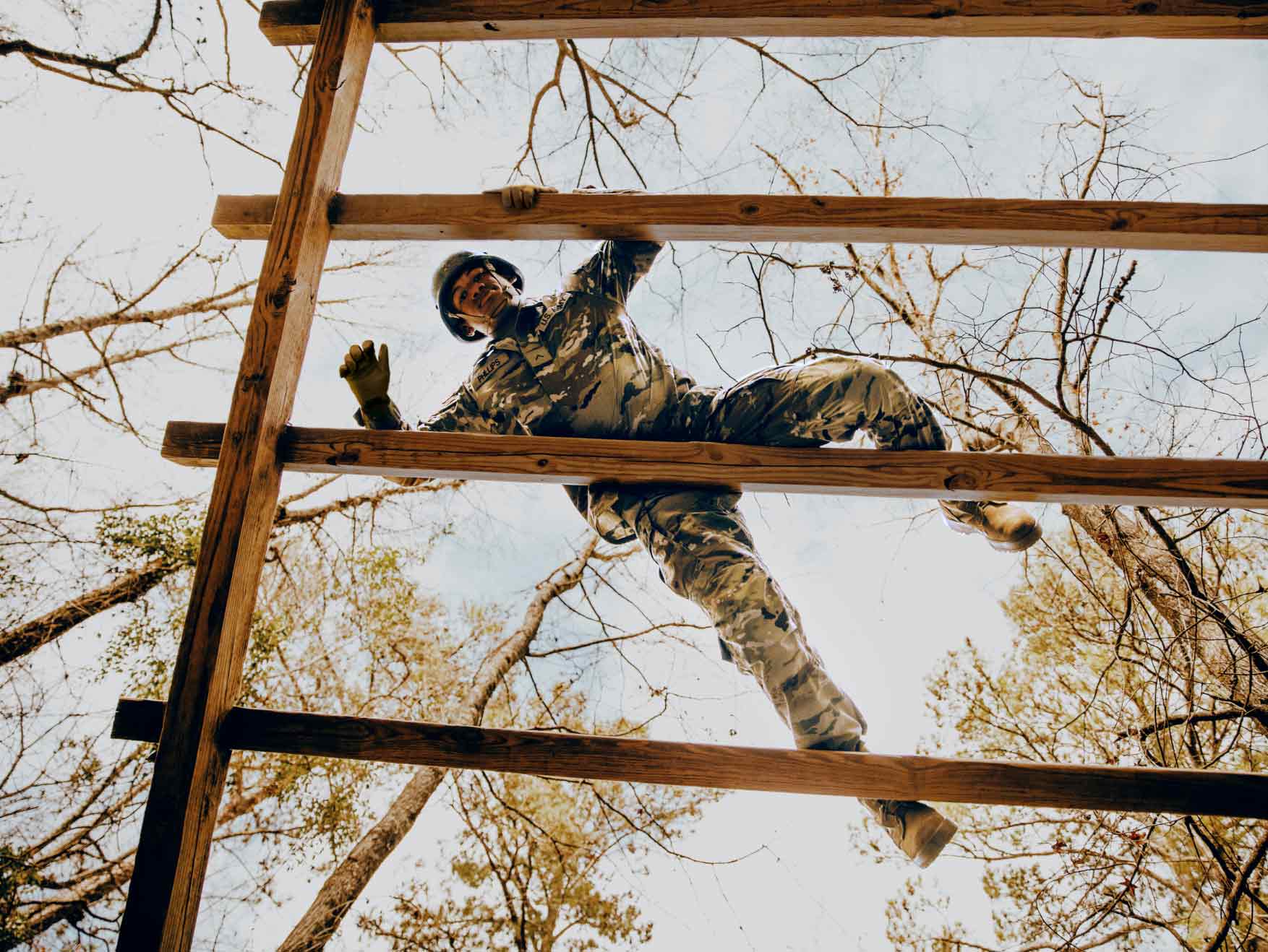 A Soldier in combat uniform climbs up a wooden obstacle on an outdoor obstacle course