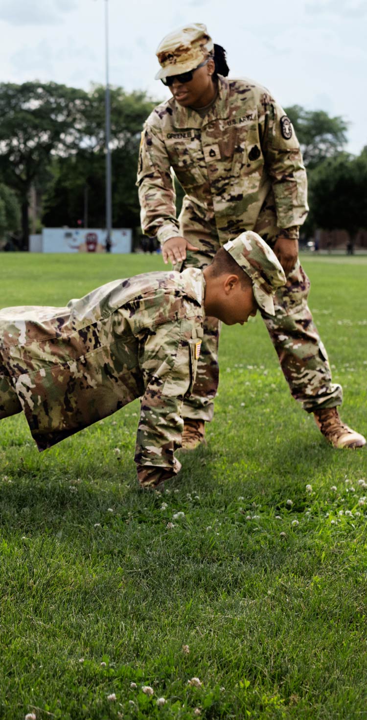 Una drill sergeant de pie al lado de un grupo de reclutas enlistados, enfocados en completar una flexión sobre el césped